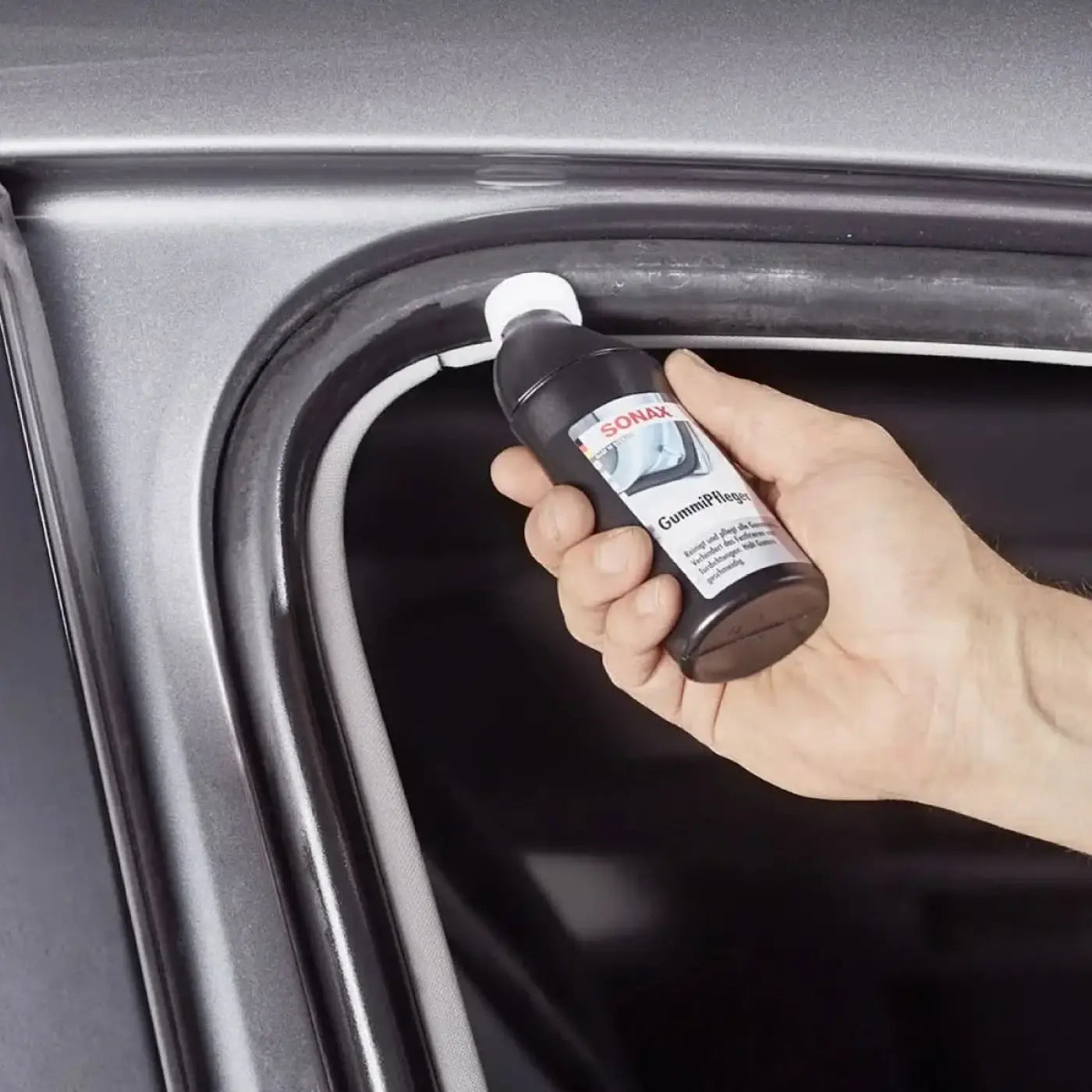 Schwarze sonax gummipfleger flasche mit weißem deckel wird an der türdichtung eines autos gehalten