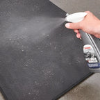 Eine hand sprüht den grauen und weißen sonax xtreme autoinnenreiniger 500ml auf eine dunkel strukturierte auto fussmatte